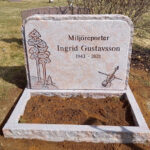 E046 i finhuggen rödflammig granit med råarbetat kantslag. Djupblästrad gravyr, dekorer med mattslipade detaljer, mattslipad blomlist med råarbetade sidytor.